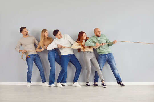 Group of happy friends playing tug of war and having fun together. Team of confident strong young adult people standing by grey color wall background, holding rope and pulling it in one direction