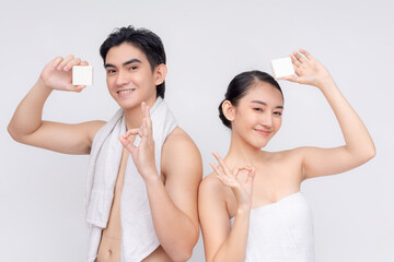 A young asian couple in their early 20s wrapped in white towels poses with a white collagen soap, making ok signs. Isolated on a white backdrop. - Powered by Adobe
