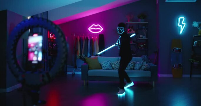 Young anime fan expresses himself on camera dim lighting and elements of blue neon lighting in his clothes make his image memorable and bright. Tiktoker performs martial arts moves in fight for likes.