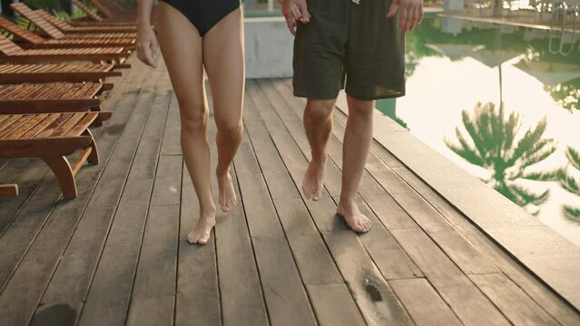 Young couple feet walk barefoot along swimming pool at tropical hotel. Boyfriend and girlfriend fit legs go at wooden deck holding hands on tropical resort hotel. Romantic vacation on beach club.