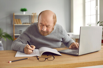 Portrait of senior man using laptop computer and making notes on paper. Serious elderly man in...