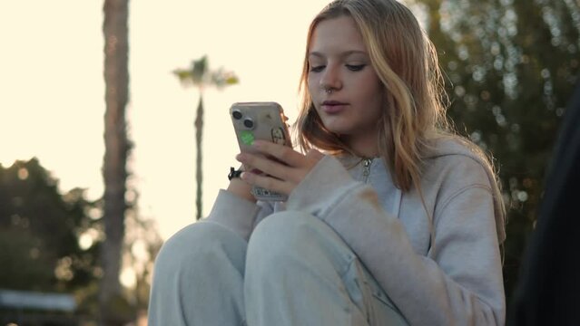 happy teenage girl with blond long hair sits in the park and watches a video or movie on the phone. High quality FullHD footage