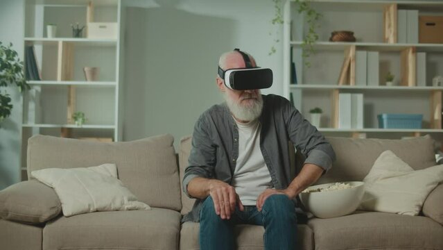 A Modern Old Man With Gray Beard in VR Glasses Examines Everything Around. An Elderly Man in VR Glasses with a Popcorn Sitting on the Sofa. VR to Help the Elderly.