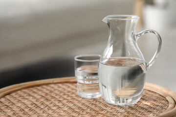 Jug and glass with clear water on wicker surface against blurred background, closeup. Space for text