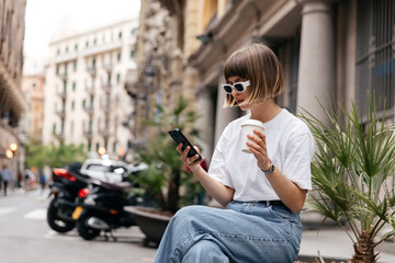 Fototapeta na wymiar Stylish modern girl with short hair is looking at smartphone and enjoying coffee outdoor on background of historical city