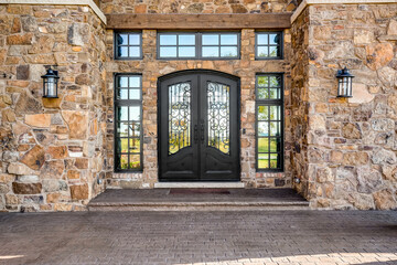 A stone sided home detail with an iron door and surrounded by blacked framed windows.