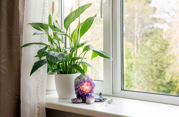 Amethyst geode lamp illuminated, spiritual calming home atmosphere. Air cleaning plant flower Spathiphyllum, spath or peace lily growing on window sill.