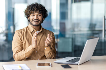 Joyful indian man in casual wear clapping hands while sitting at workplace with laptop in modern...