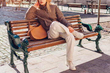 Fashionable woman sitting on bench on street of European city wearing stylish pants, boots and jacket. Spring outfit