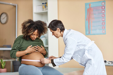 Smiling pregnant Black woman sitting on examination table in medical center while mature Caucasian female gynecologist checking abdominal girth with measuring tape - obrazy, fototapety, plakaty