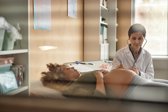 Look through ward window of friendly mature Caucasian gynecologist with stethoscope checking fetal heart tones while pregnant Black female patient lying down during clinic visit