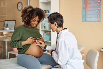 Smiling young African American expectant mother sitting on medical table in clinic while female...