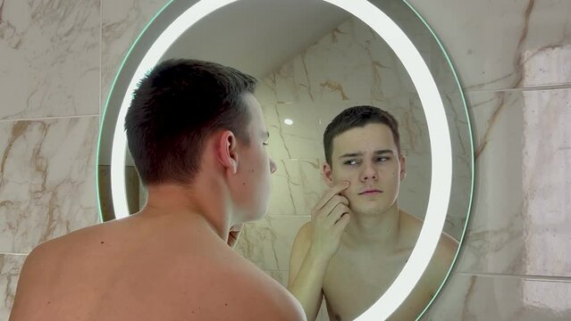 Unhappy teenager looks at his pimples in the mirror. 