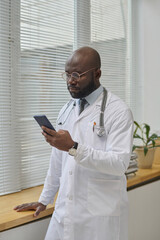 Young african american doctor in uniform standing by window in his office and browsing smartphone