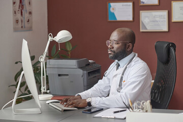African american doctor in white lab coat sitting at his desk in office and working on computer