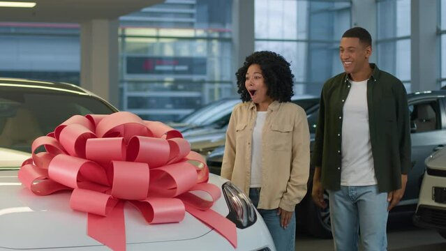 African American couple boyfriend girlfriend in dealership showroom surprise new crossover car gift birthday present with red bow on auto happy woman wife rejoice enjoy luxury automobile hug husband