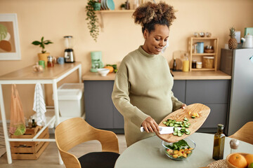 High angle shot of smiling Black expectant mother with kitchen knife adding freshly cut cucumber to...