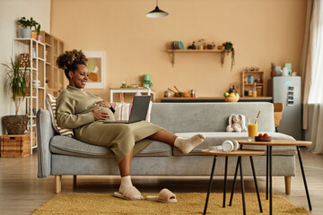 Wide shot of happy pregnant African American woman lying back on big couch in living room with...
