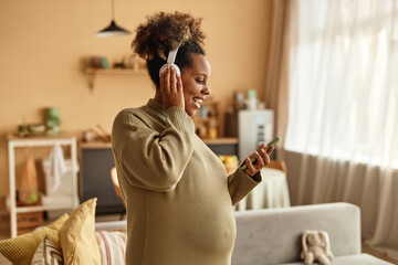 Waist up shot of smiling pregnant Black woman vibing to music looking at smartphone in hand while standing in cozy living room