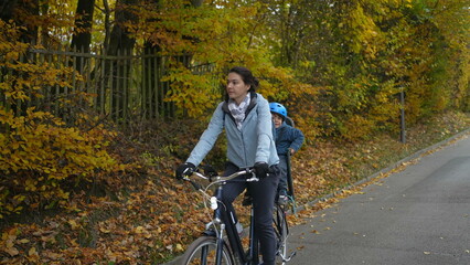 Fototapeta premium Mother rides bicycle with child in back seat during autumn fall day. Parent riding bike with son wearing helmet and jacket