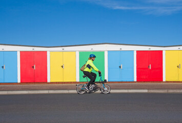Cycling image of a lady on a bicycle riding past a row of brightly coloured beach huts. Good copy...