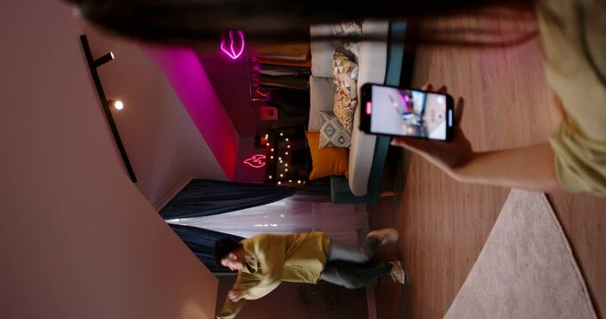 Girl enthusiastically looks screen of her mobile phone, she records video of boyfriend vigorously dancing to fashionable track, after they edit reel, they will post it on their channel. Vertical video