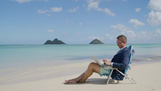 A businessman in a suit is focused on his laptop while seated on a beach chair, with the tranquil ocean and distant islands framing him. Slow Motion, 4K Hi Quality. 