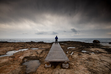 Man standing alone at the edge of rocky beach in Stormy Weather. Cape Greko Cyprus.