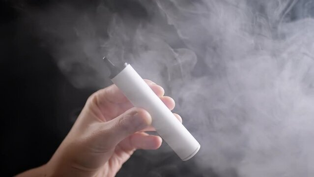 Close up, Smoking a Vape or E-cigarette in a Dark Room on a Black  Background. A hand holding vape. Blowing thick clouds of white smoke.  Texture. Filling smoke. Smoking cigarettes. Bad habit. Nicotine.