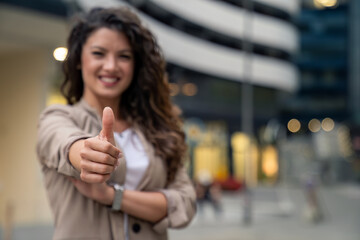 Smiling confident female business professional standing outside an office building looking at camera showing thumbs up as sign of sign of success. Focus is on the hand.