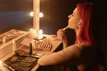 Side view of red-haired Caucasian girl with tattoo applying makeup and putting on face powder with...