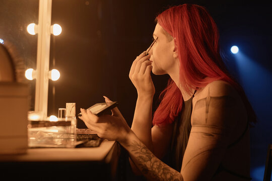 Profile view of of tattooed young woman putting on makeup and applying eyeshadow with brush in dressing room before dance show