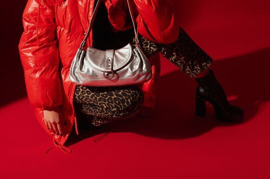 Trendy metallic leather baguette bag, purse in fashionable outfit. Woman wearing red jacket, leopard print jeans, platform boots, posing on red backdrop. Copy, empty, blank space for text