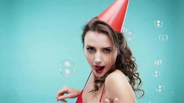 Woman, dance and celebrate in studio with bubbles for party, winning and come join me on new year. Excited Face of person, winner or model with energy, happy and portrait in hat on a blue background