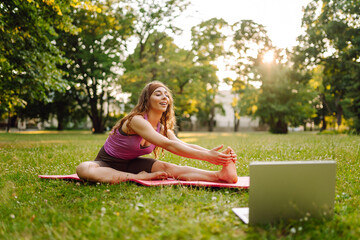 Young woman doing fitness stretching yoga exercise in the park while using laptop for online class or virtual tutorials. Training workout. Healthy lifestyle, sport. Online training, course.