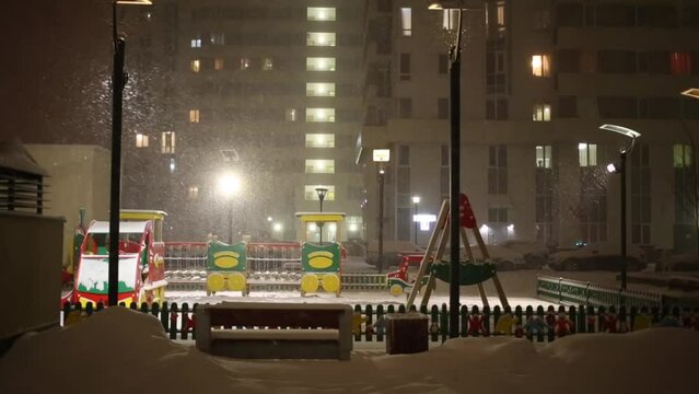 Snow falling on empty playground near apartment building at night.