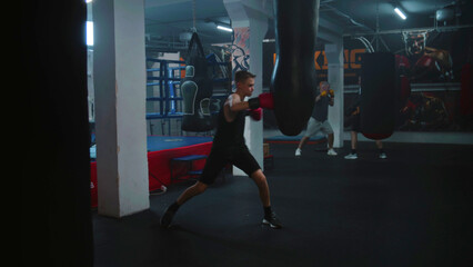 Focused young fighter listens coach command and starts hitting punching bag in dark boxing gym....