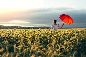 Junge Frau mit einem roten Schirm steht in einem Getreidefeld und schaut Richtung Sonnenuntergang. Ein Kirchturm am Horizont. - obrazy, fototapety, plakaty