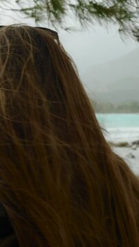 Vertical video. Alone on an empty seaside in a cool day, a woman shelters under the pine branches and admires the turquoise water in the bay against the backdrop of mountains. Feminist concept, feelin