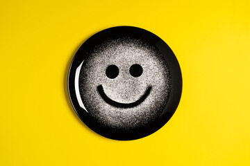 Smiling face, concept made with plate and flour, yellow background, black plate, happy mood