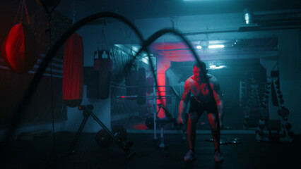 Professional boxer does cardio or endurance workout before championship fight. Athletic man exercises with battle ropes in dark boxing gym with LED lighting. Physical activity and CrossFit training.