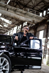 Man in black leather jacket stands near black car in abandoned building.