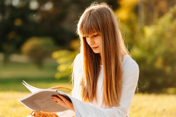 Naklejka premium Concentrated young woman with bangs is reading a book while sitting in park studying for an exam.