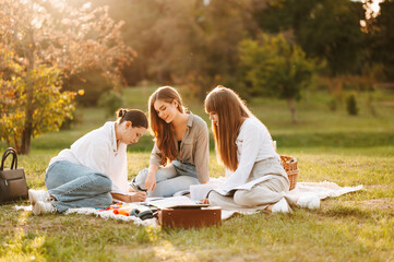 Three cheerful young female students organised a learning picnic together in park.