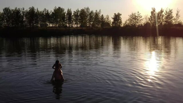 Girl sitting on boys shoulders in water of lake at sunset, mobile phone video.