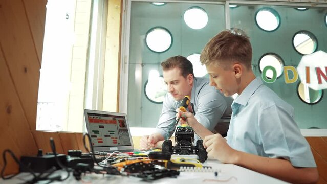 Caucasian teacher pointing at laptop screen and giving advise for programming system or coding prompt while smart high school boy listen and use equipment to repair robot at STEM classroom