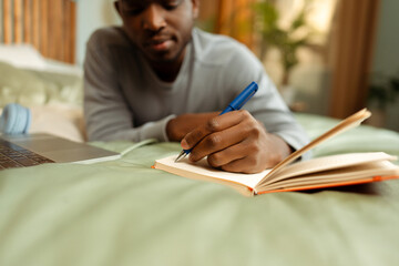 Portrait of attractive African American student lying on bed learning language, taking notes