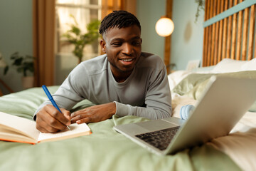 Smiling attractive African American man, student using laptop, taking notes, preparing for exam