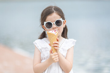 Beautiful little girl in sunglasses and white dress eating an ice cream. High quality photo