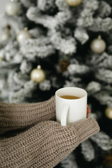 Close up of person's hands in warm sweater holding a white mug with hot coffee in front of decorated Christmas tree
